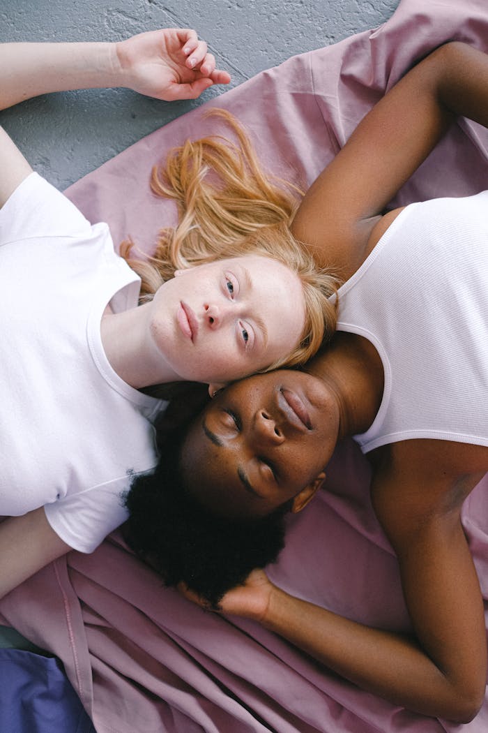 Two women, an interracial couple, lying peacefully on pink fabric, expressing love and connection.